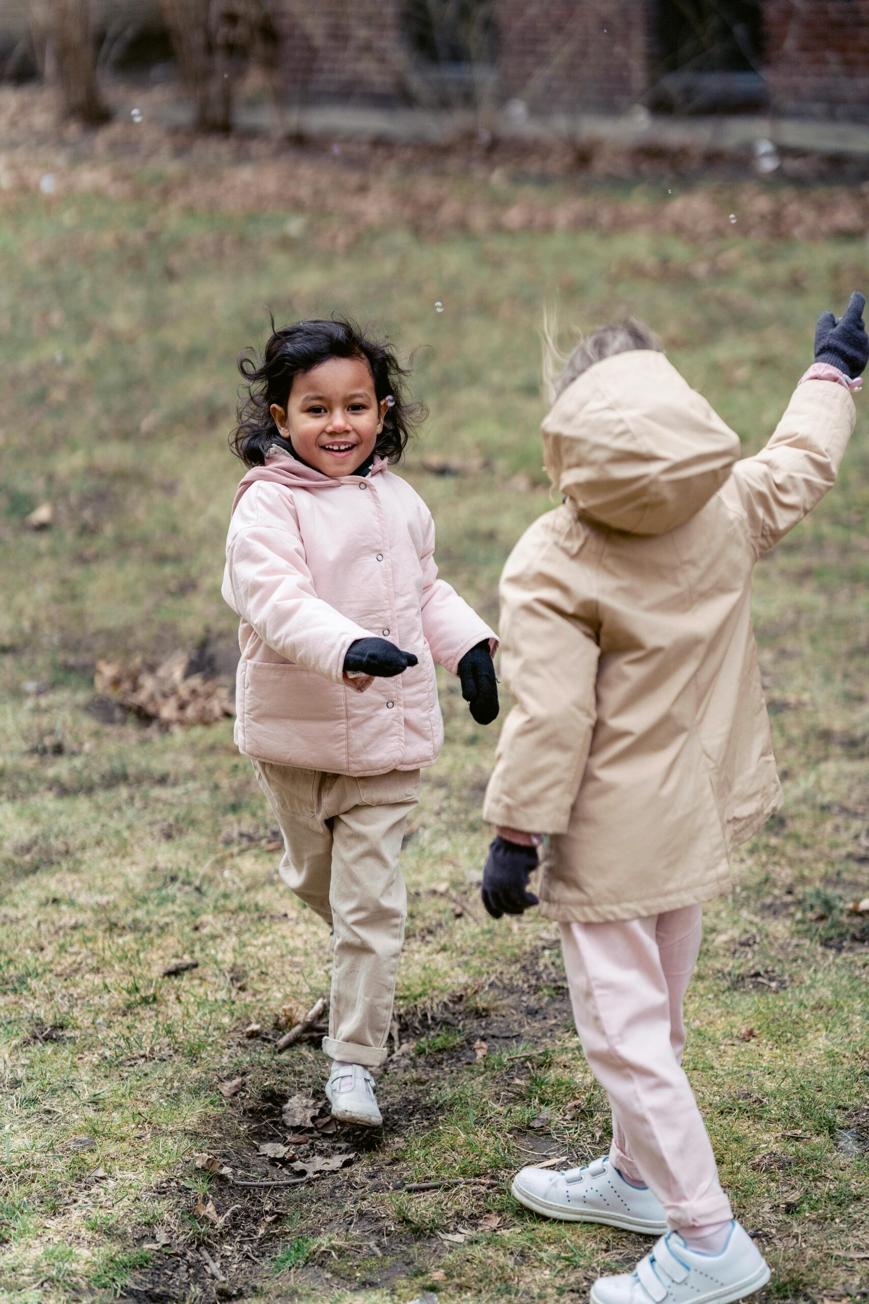 Smiling ethnic child looking forward while running against unrecognizable girlfriend in warm clothes on meadow under soap bubbles